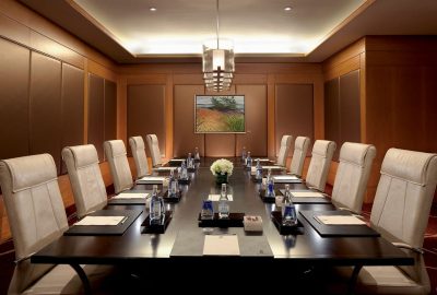 Long dark wood conference table with white leather chairs, water bottles, and white flowers in a wood-paneled room.