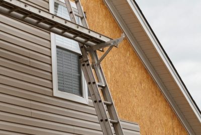 Extension ladder leaning against a house with beige siding and exposed OSB sheathing.