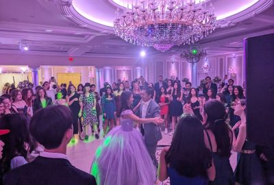 A young woman in a lavender gown dances with a partner in a suit, surrounded by guests in a ballroom under a chandelier.