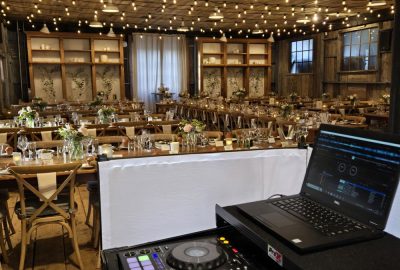 DJ setup with a laptop and controller overlooking a rustic banquet hall decorated with string lights and floral centerpieces.