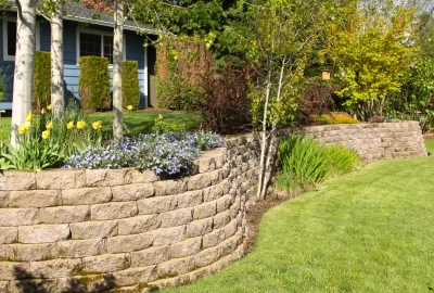 Curved stone retaining wall with yellow and blue flowers bordering a green lawn in a landscaped yard.