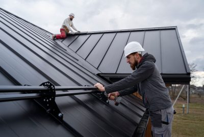 A construction worker in a white hard hat uses a power drill to install metal rails on a dark metal roof.