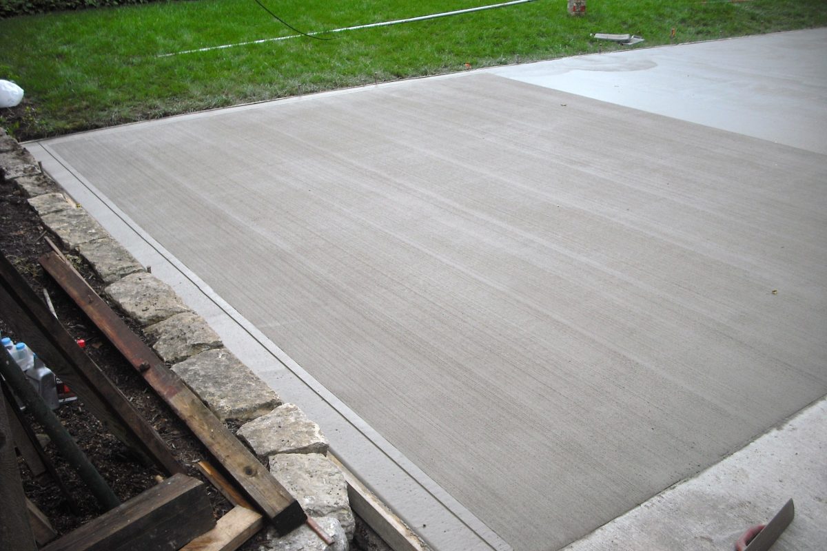 Newly poured concrete surface with a broom finish, adjacent to a grassy lawn and stone edging.