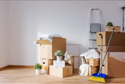 Packed cardboard boxes, a broom, and a step ladder in an empty room with white walls and wooden floors.