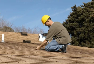 Roof inspector in a yellow hard hat kneeling on a shingle roof and taking notes on a clipboard.