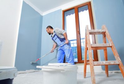 A man in blue overalls uses a paint roller to paint a light blue wall in a room with a window and a ladder.