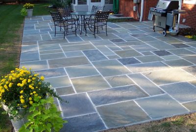 A stone patio with a dining set, a grill, and yellow flowers in a pot in the foreground.