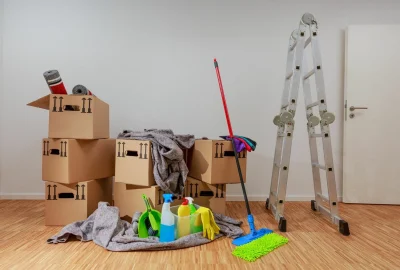 Cardboard boxes, cleaning supplies, and a silver ladder on a wooden floor in a room.