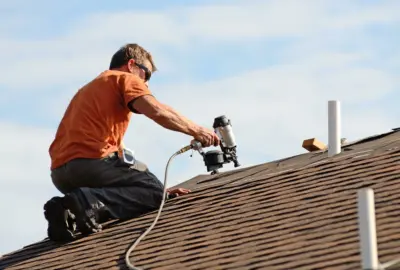 Roofer in an orange shirt using a pneumatic nail gun to install shingles on a roof.