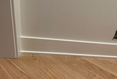 Close-up of white baseboard trim between a white wall and light wood flooring.