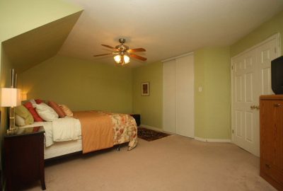 Bedroom with light green walls, a bed with an orange floral comforter, a ceiling fan, and beige carpeting.