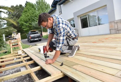 Man using a power drill to install wooden boards on a deck under construction.