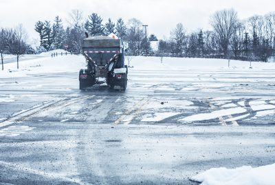 A salt spreader truck drives across a snow-covered asphalt lot on a winter day.