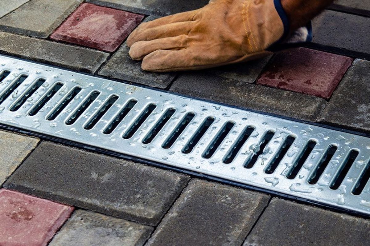 Gloved hand installing a metal drainage grate into a brick paved walkway.
