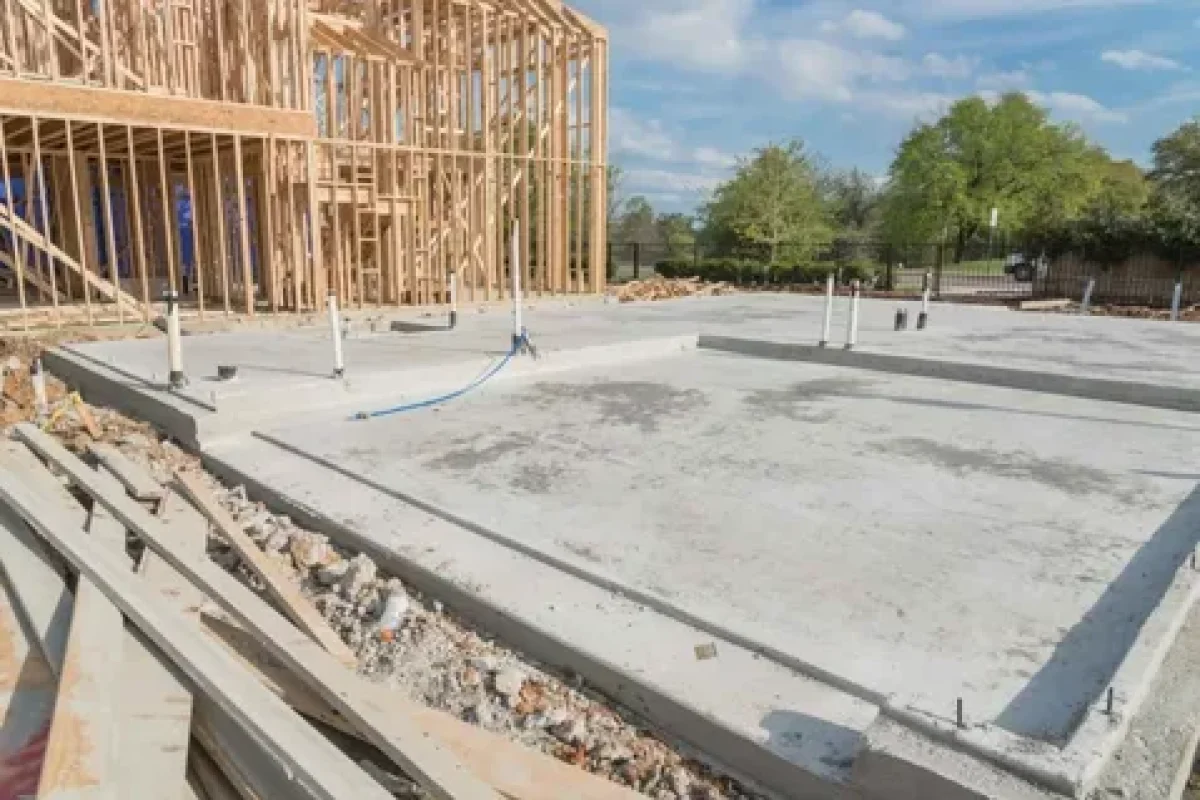 Concrete foundation slab with plumbing pipes and a wooden house frame under construction.