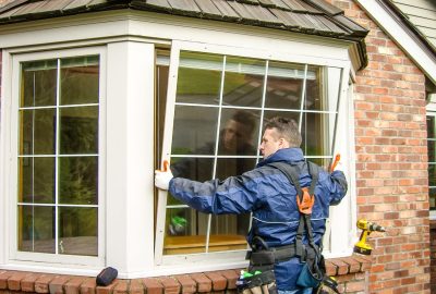 A worker in a blue jacket and tool belt installs a white bay window on a brick house.