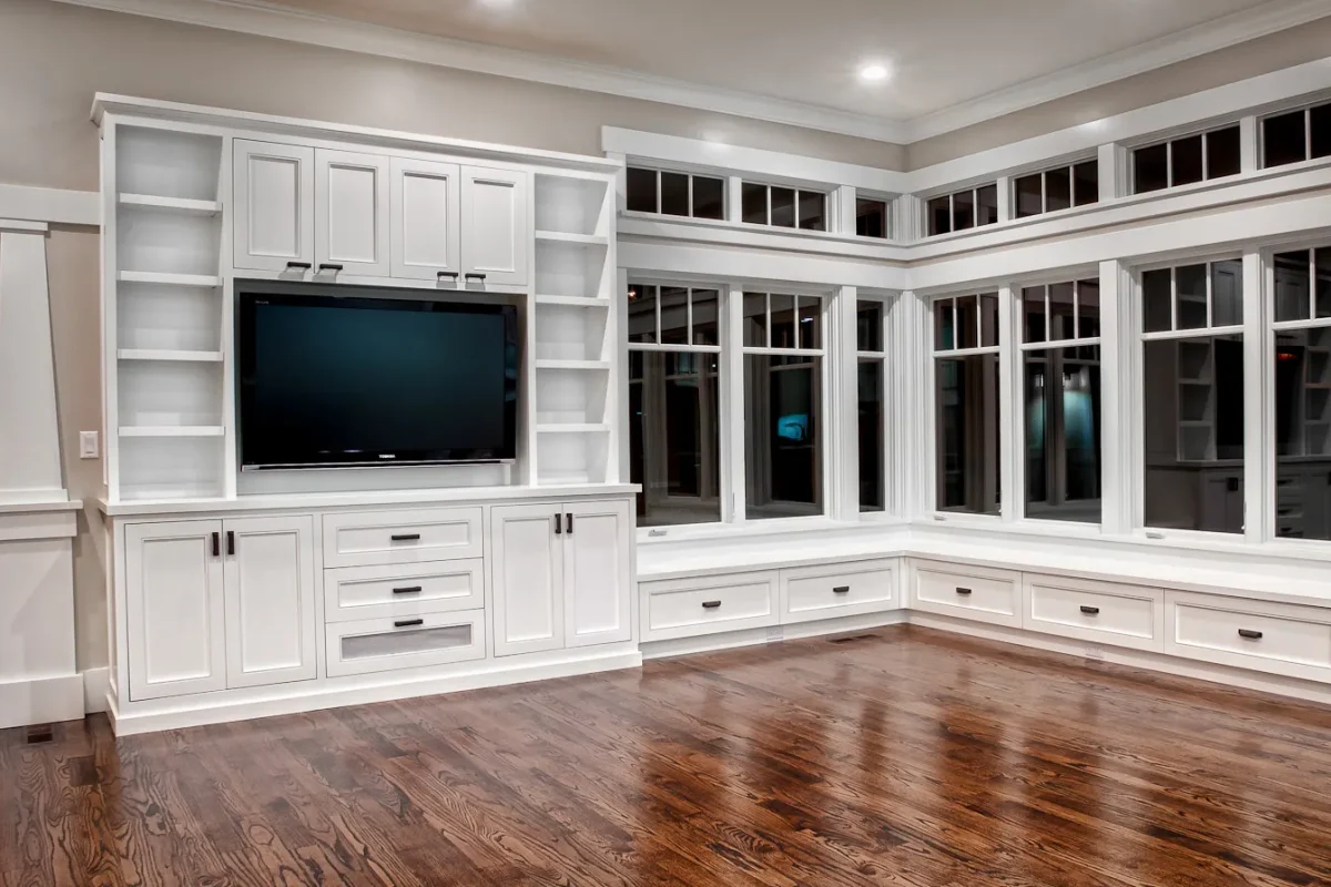 White built-in entertainment center and window bench with storage drawers on dark hardwood floors.
