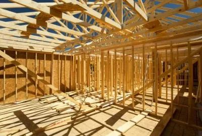 Wooden wall framing and roof trusses of a house under construction under a clear blue sky.