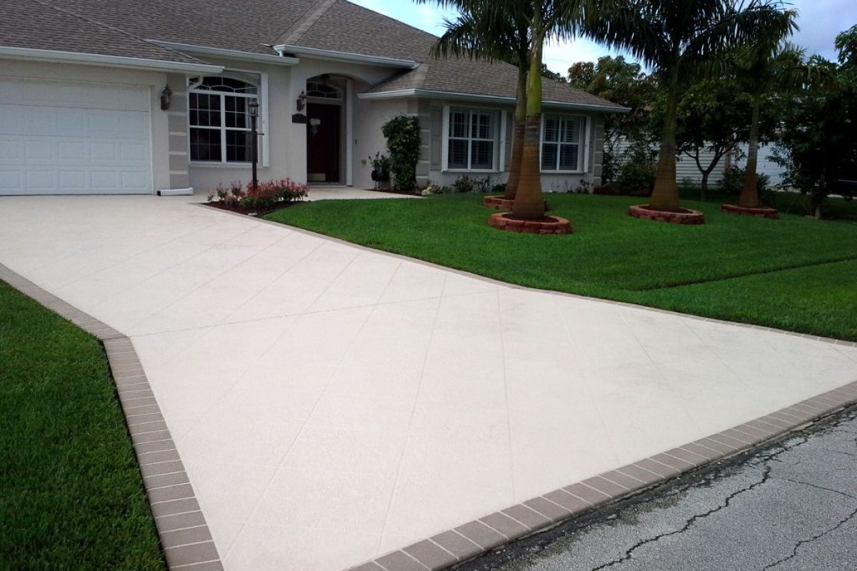 Light beige stamped concrete driveway with a brick border in front of a suburban home with palm trees.