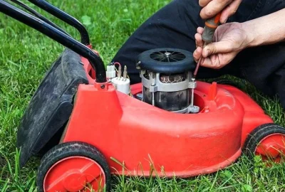 Person repairing a red electric lawnmower with a screwdriver on a lawn.