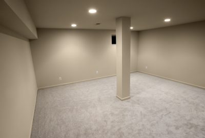 Empty room with beige walls, light-colored carpeting, and a central support pillar under recessed lighting.