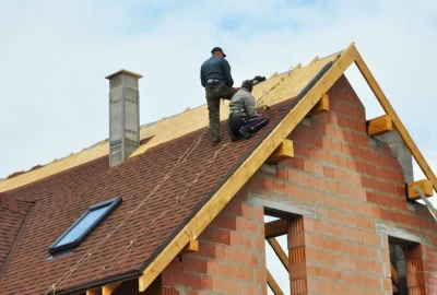 Two construction workers installing brown shingles on the roof of a brick house under a clear sky.