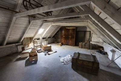 Dusty attic with white sloped ceilings, scattered boxes, wooden crates, and a large wooden wardrobe.