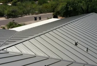 Gray standing-seam metal roof with ridges and vents in a residential area.