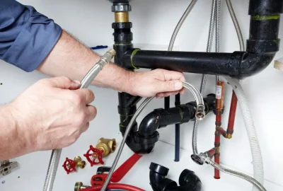 Plumber's hands connecting a flexible braided water supply hose to pipes under a kitchen sink.