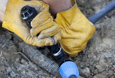 Hands in yellow work gloves tightening a plastic pipe fitting in the dirt.