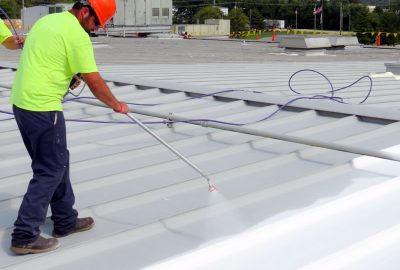 Worker in a neon shirt and orange hard hat spraying a white coating onto a corrugated metal roof.