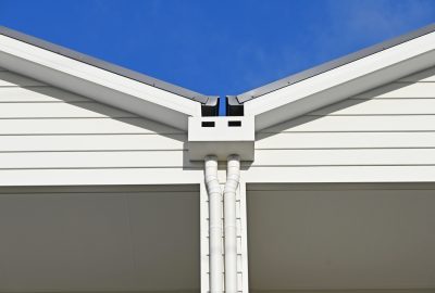Close-up of a white gutter valley and twin downspouts on a white house against a clear blue sky.