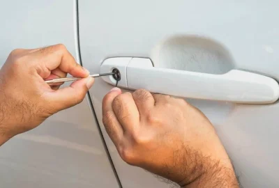 Hands using lock picking tools on a white car door handle.