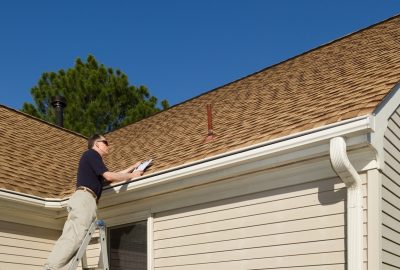 A man on a ladder inspecting the roof and gutters of a house while holding a clipboard.