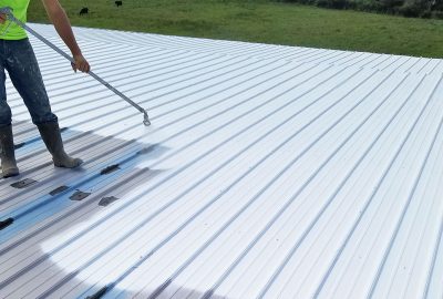 Worker using a spray gun to apply white coating to a corrugated metal roof.