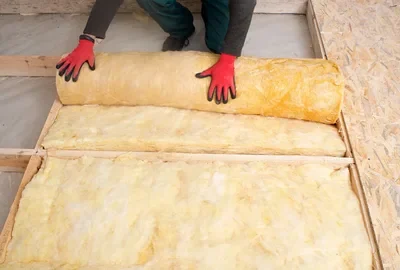 Worker in red gloves unrolling yellow fiberglass insulation between wooden floor joists.