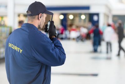 Security guard in a blue jacket and cap using a handheld radio in a blurred public area.