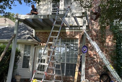 Man working on a wooden deck frame attached to a brick house with extension ladders positioned nearby.