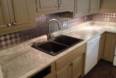 Kitchen with tan cabinets, light countertop, dark double sink, and a partially installed silver patterned backsplash.