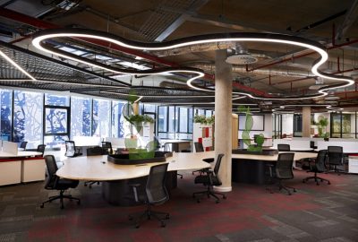 Modern industrial office with curved wooden desks, ergonomic black chairs, and flowing LED ceiling lights.