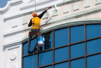 Painter suspended by ropes using a roller to apply white paint to the exterior facade of a building.