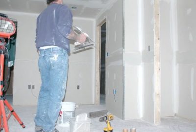 A construction worker applies drywall compound to walls in a room under renovation.