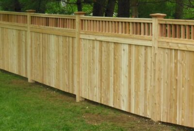 Light wood privacy fence with a decorative slatted top section, set on a green lawn against a backdrop of trees.