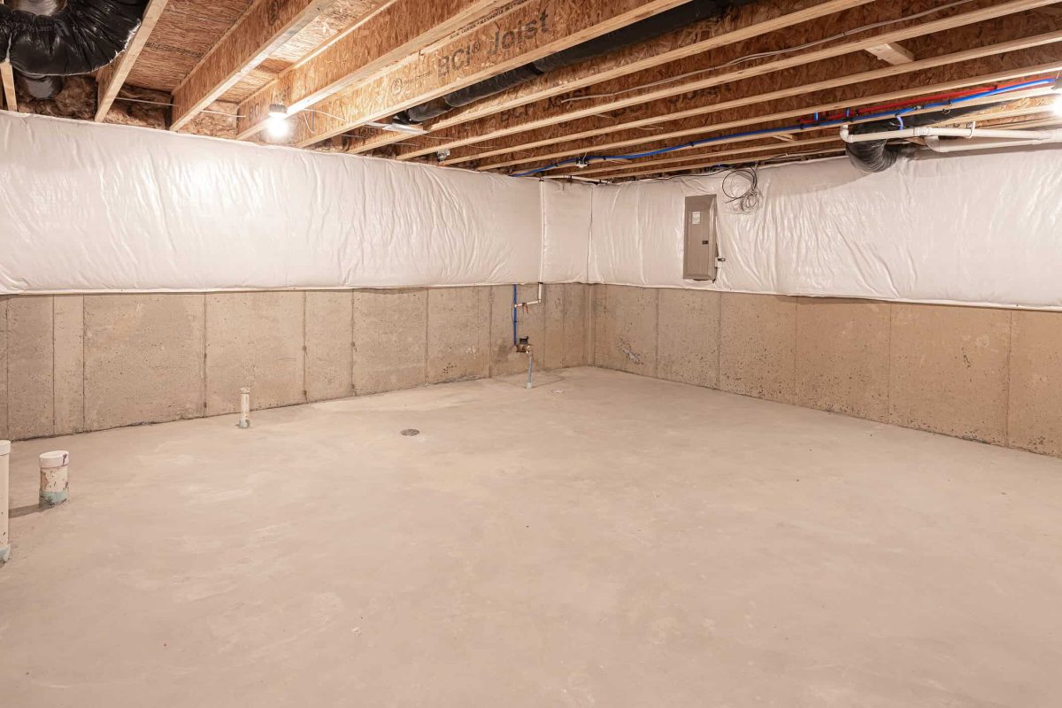Unfinished basement with concrete floors, insulated walls, and exposed wooden ceiling joists with piping.