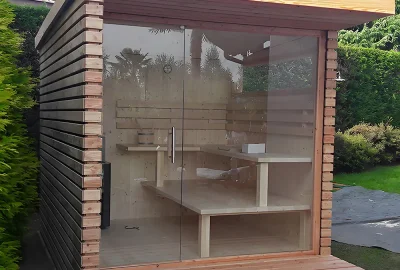 Modern wooden outdoor sauna with a glass front door and tiered seating, situated in a garden.