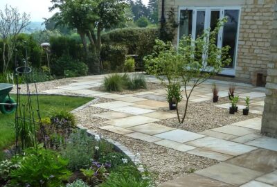 Stone patio with light pavers and gravel, potted plants, and a small tree next to a stone house.