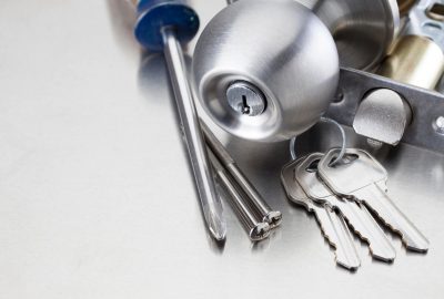 Silver doorknob, a set of keys, a screwdriver, and screws on a metallic surface.