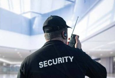Rear view of a security guard in a black uniform and cap, using a walkie-talkie in a bright building.