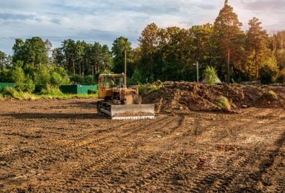 Yellow bulldozer on a dirt construction site with tire tracks and a large mound of earth near a forest.