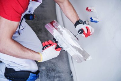 A person in white overalls and red-and-white gloves uses a large putty knife to apply white plaster to a wall.
